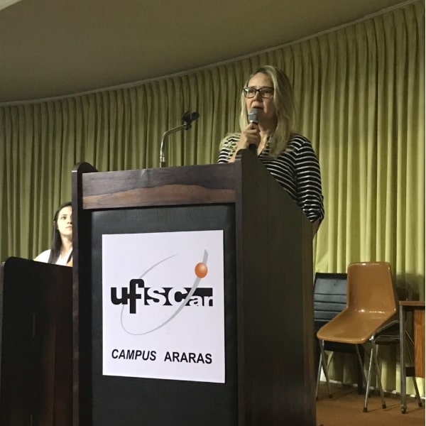 Fotografia da professora Dra. Monalisa Sampaio Carneiro no palco do auditório do Centro de Ciências Agrárias segurando um microfone, apresentando o programa de pós-graduação e fazendo discurso de abertura do III Simpósio de Produção Vegetal e Bioprocessos Associados. Ela se encontra atrás do púlpito de madeira que possui impresso na parte da frente o símbolo da Universidade Federal de São Carlos com o escrito: campus Araras. No canto esquerdo da foto encontra-se Juliana Uzan, presidente da comissão do Simpósio sentada na ponta da mesa principal.