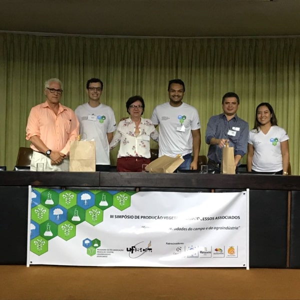 Fotografia dos palestrantes que participaram da mesa redonda “Aproveitamento de resíduos agroindustriais” junto com a moderadora Profa. Dra. Sandra Ceccato Antonini e discentes da comissão organizadora do simpósio que entregaram um brinde a cada um dos palestrantes. Encontram-se em pé no palco do auditório, atrás da mesa principal na seguinte ordem: Prof. Dr. Alcides Leão, Francisco José da Silva Neto, Profa. Dra. Sandra Ceccato Antonini, José Machado Neto, Prof. Dr. Severino de Alencar e Dra. Ligianne Shirahigue Viani.