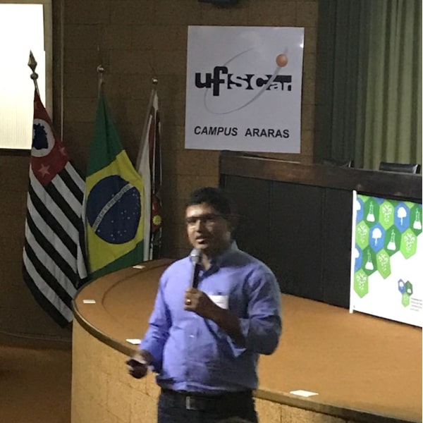 Fotografia do palestrante Dr. Renato de Carvalho no centro da imagem. Ele se encontra à frente do palco do auditório, segurando um microfone e um pointer. Ao fundo da imagem tem-se o palco com a mesa principal à direta, um cartaz contendo o símbolo da Universidade Federal de São Carlos - campus Araras ao centro, e à esquerda 3 bandeiras hasteadas: do estado de São Paulo, do Brasil e do município de Araras.