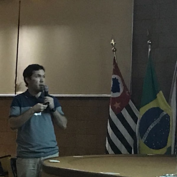 Fotografia do palestrante Dr. Gabriel Moura Mascarin segurando um microfone e apontando para sua apresentação na tela branca de projeção que se encontra no centro do palco do auditório do Centro de Ciências Agrárias. À direita da imagem estão as bandeiras do estado de São Paulo e do Brasil hasteadas no fundo do palco.