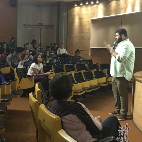 Fotografia do palestrante Eng. Agr. Lucas Cirilo à direita da imagem voltado para a plateia que se encontra à esquerda da imagem. Ele segura o microfone e gesticula para os ouvintes sentados nas poltronas do auditório. Dentre os ouvintes tem-se alunos e professores.