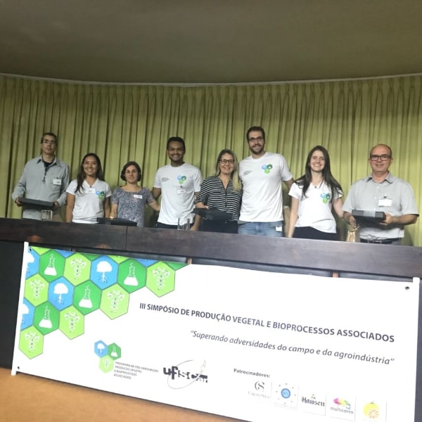 Fotografia dos palestrantes que participaram da mesa redonda “Adaptação às mudanças climáticas na agricultura” junto com a moderadora Profa. Dra. Monalisa Sampaio Carneiro e discentes da comissão organizadora do simpósio que entregaram um brinde a cada um dos palestrantes. Encontram-se em pé no palco do auditório, atrás da mesa principal na seguinte ordem: Dr. Alexandre Ortega Gonçalves, Dra. Ligianne Shirahigue Viani, Profa. Dra. Sara Andrade, José Machado Neto, Profa. Dra. Monalisa Sampaio Carneiro, Rafael Kupper, Bruna Bettini e Prof. Dr. Marcelo Menossi Teixeira. À frente da mesa principal tem-se uma faixa impressa com o logotipo e o título do simpósio, e os logotipos dos patrocinadores.