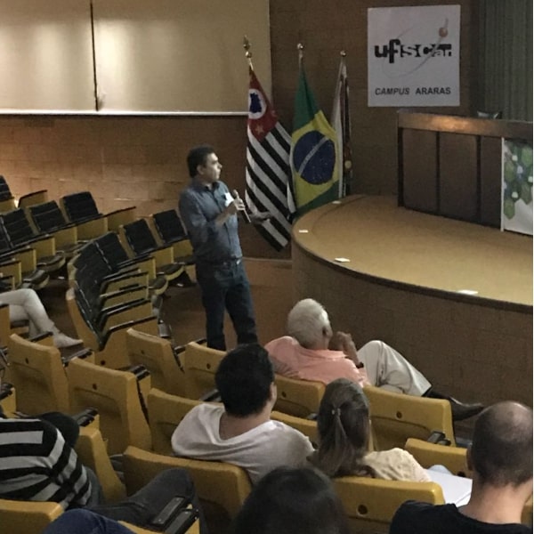 Fotografia do palestrante Prof. Dr. Severino de Alencar durante sua apresentação, voltado e apontando para a tela branca de projeção e segurando um microfone. O cenário atrás do palestrante é composto do palco com a mesa principal e ao fundo um cartaz contendo o símbolo da Universidade Federal de São Carlos - campus Araras e 3 bandeiras hasteadas: do estado de São Paulo, do Brasil e do município de Araras. A porção inferior da imagem mostra a plateia, com os participantes olhando para a tela no palco.