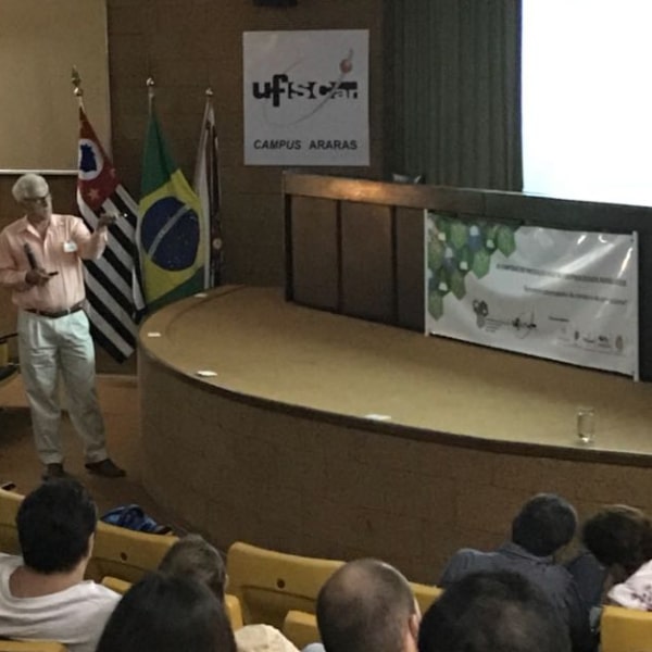 Fotografia do palestrante Prof. Dr. Alcides Leão durante sua apresentação, voltado e apontando para a tela branca de projeção e segurando um microfone. O cenário atrás do palestrante é composto do palco com a mesa principal e ao fundo um cartaz contendo o símbolo da Universidade Federal de São Carlos - campus Araras e 3 bandeiras hasteadas: do estado de São Paulo, do Brasil e do município de Araras. A porção inferior da imagem mostra a plateia, com os participantes olhando para a tela no palco.